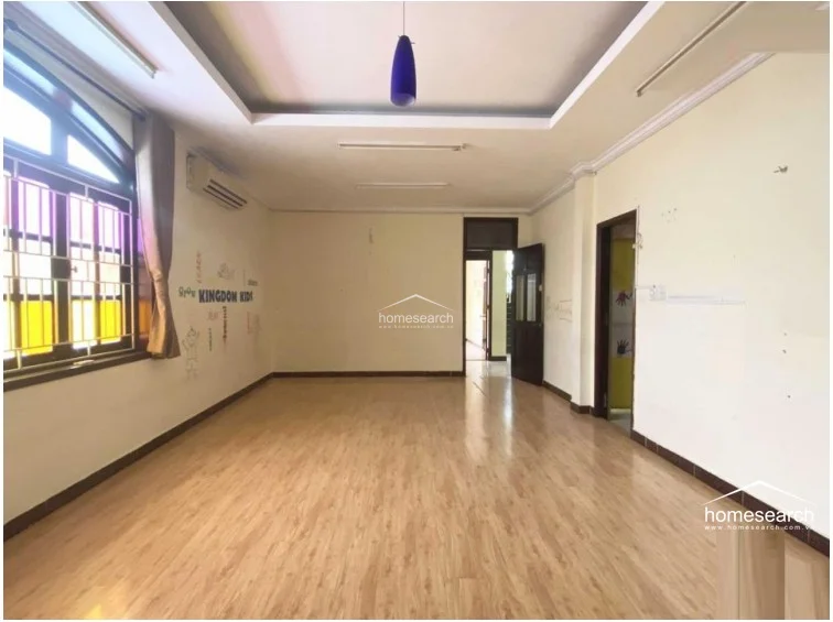 This bright, multi-purpose room features warm wood-look laminate flooring and a recessed ceiling with a modern pendant light. The space is equipped with a wall-mounted AC unit and large windows with decorative grilles, allowing for ample natural light, while the walls retain some playful children's wall decals.