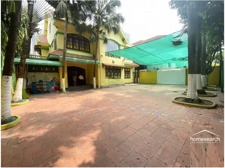 This image reveals the side courtyard of a yellow-painted building, which appears to be utilized as a play area or outdoor recreational space. A large green shade canopy covers the paved terrace, and the area is bordered by palm trees and a perimeter fence, indicating a functional, family-oriented outdoor space.