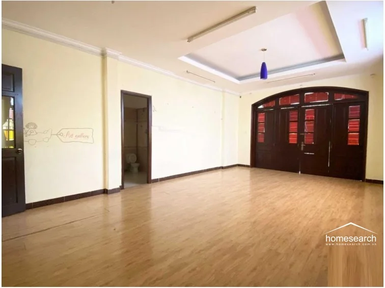 This expansive room serves as a versatile living or activity area, anchored by a striking set of dark wood arched double doors that add a touch of architectural character. The space features light-toned wood flooring, a recessed ceiling design, and a direct view into an adjacent bathroom, reflecting a well-maintained, neutral interior.