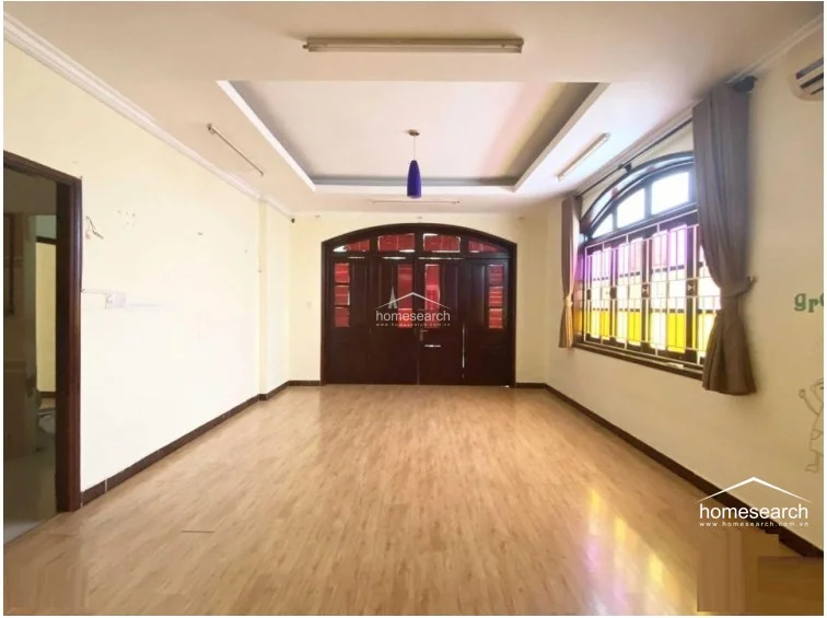 This view highlights the room's open layout, showcasing the contrast between the light wood-look flooring and the dark, ornate wooden entrance doors. The room benefits from significant natural light streaming through large, arched-top windows, and the overall condition appears clean and ready for flexible use as a living area or workspace.