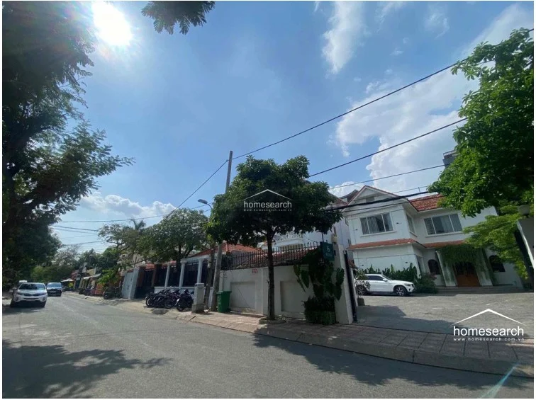 A wider street-level view captures the property's corner positioning and its integration into a quiet, tree-lined neighborhood. The white perimeter wall and gated access provide security, while the surrounding foliage contributes to a serene, suburban atmosphere.