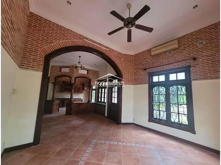 This interior view shows a spacious living area characterized by high ceilings, exposed brick upper walls, and a large, elegant wooden archway that connects to the kitchen. The room features terracotta tile flooring, a ceiling fan, and a wall-mounted AC unit, with dark wood-framed windows fitted with decorative security grilles. The layout offers an open, flowing design, though the interior finishes appear dated and in need of refreshing.
