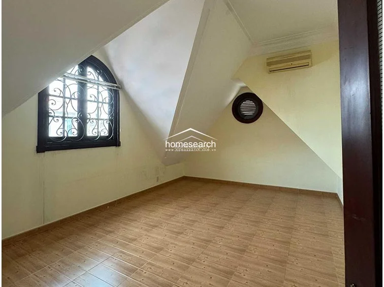 This attic-style bedroom features a unique slanted ceiling design that creates a cozy, architectural aesthetic. The room is finished with light-toned wood-look tile flooring and pale yellow walls, complemented by a wall-mounted air conditioning unit and a distinctive arched window with decorative iron security bars.