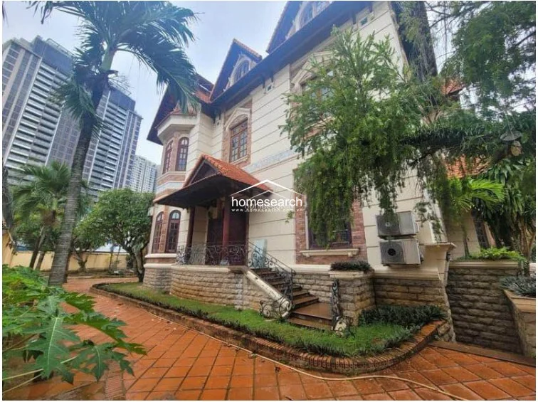 This view highlights the side elevation of the villa, showcasing its classic architectural details, including arched windows and a covered porch with decorative iron railings. The exterior walls utilize a combination of smooth stucco and textured stone base finishes, while two large AC units are mounted prominently on the wall. The landscaping is lush but appears slightly overgrown, indicating that the property could benefit from professional exterior upkeep.