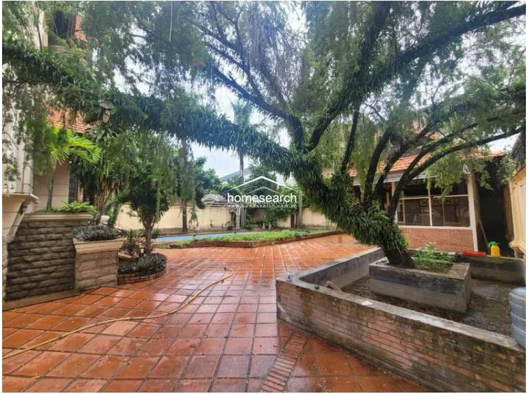 This outdoor area features a spacious terracotta-tiled patio surrounding a swimming pool, set against a backdrop of mature trees and a stone-clad exterior wall. The space is well-defined by brick-edged planters, offering a private, garden-like atmosphere despite the urban setting.