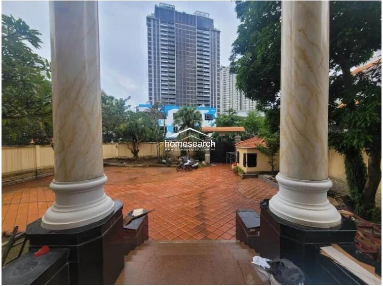 This grand entrance features a sweeping staircase leading down to a paved courtyard, framed by two prominent marble-style columns. The property offers a clear view of a modern high-rise in the distance, contrasting the classic architectural elements with the surrounding urban landscape.