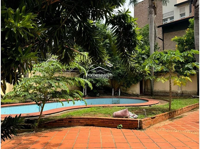 This shot features a curved, in-ground swimming pool situated within a private garden area, framed by tropical greenery and palm trees. The pool area is paved with matching terracotta tiles, though the pool itself appears to be currently drained or in need of cleaning and maintenance. The surrounding wall provides significant privacy, though the overall space requires landscaping attention to restore its appeal.