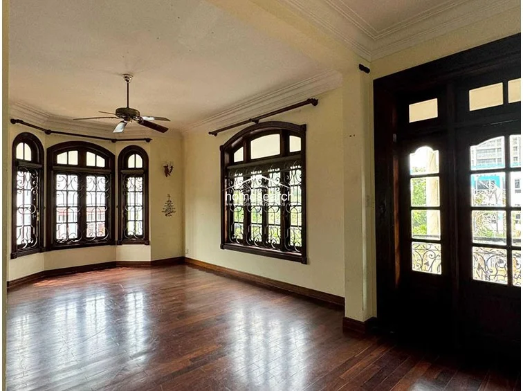 This living area showcases elegant dark hardwood flooring and intricate crown molding, complemented by a classic ceiling fan. The room is flooded with natural light from multiple arched windows featuring ornate wrought-iron security grilles, creating a bright and sophisticated atmosphere.