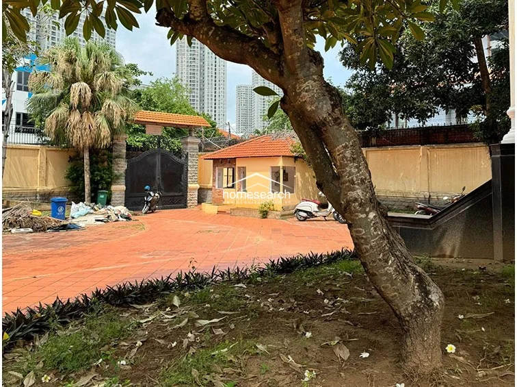 This image captures the gated entrance and security guard house, which features a matching terracotta tile roof and light-colored stucco walls. The large, ornate black metal gate provides secure access to the expansive courtyard, while the area is shaded by a mature tree. The presence of debris near the gate suggests that the property is currently unoccupied or undergoing maintenance.
