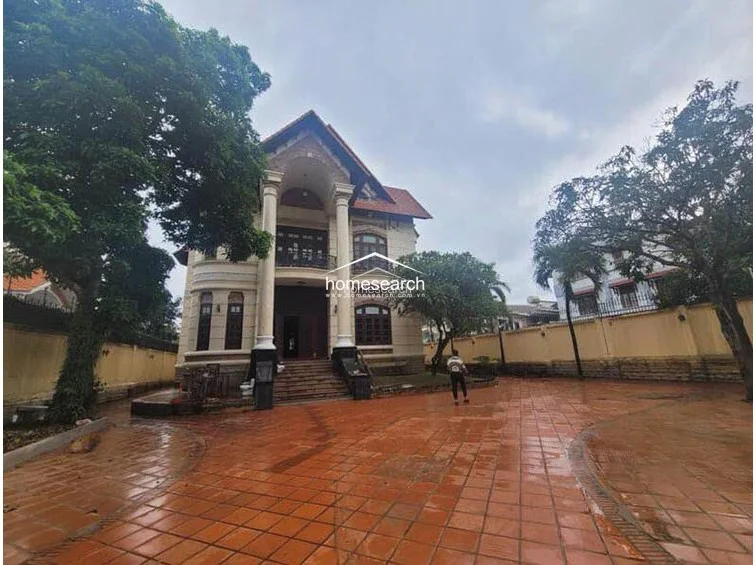 This image shows the exterior facade of a large, neoclassical-style villa featuring prominent white columns and a grand staircase leading to the main entrance. The property is surrounded by a spacious courtyard paved with terracotta tiles and mature trees, suggesting a high-end, private residential setting. The overall condition appears to be in need of some exterior maintenance, particularly regarding the grounds and masonry.