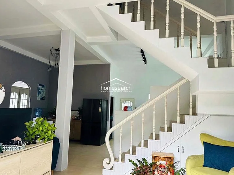 The interior living area features a clean, modern aesthetic with white walls and a striking staircase with white wooden balustrades. The open floor plan flows into a dining or kitchen area, highlighted by a large black refrigerator and a cozy seating area tucked under the stairs.