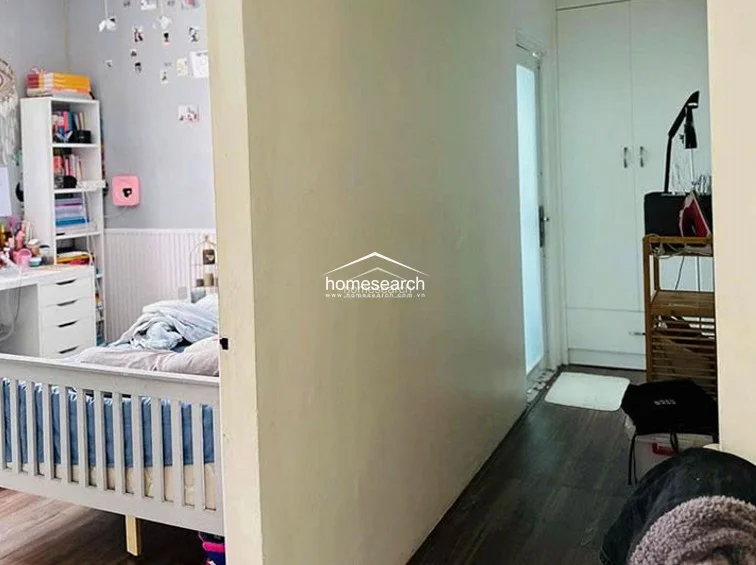 This image shows a bedroom layout featuring a light grey bed frame, a white desk with integrated shelving, and dark wood-look flooring. The room includes a built-in white wardrobe in the adjacent hallway area, maximizing storage, and is finished with neutral grey walls and wainscoting.