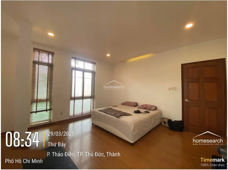 This bedroom is characterized by warm wood flooring and minimalist white walls, creating a clean and restful environment. It features tall, narrow windows fitted with wooden blinds for privacy and light control, and includes a solid wood door that adds a touch of traditional warmth to the space.