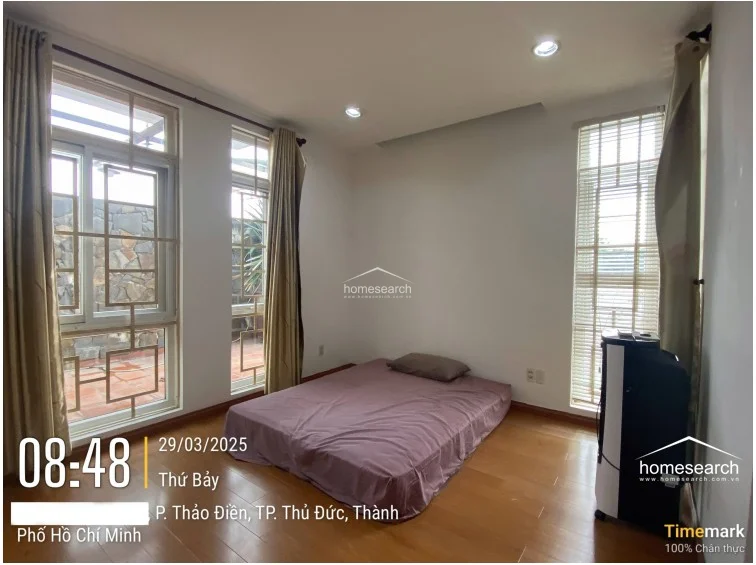 This bedroom features warm wood-tone flooring and crisp white walls, creating a clean and minimalist aesthetic. Large windows with beige curtains allow for ample natural light and provide a view of an outdoor terrace area, while a portable air cooler serves as the primary climate control fixture.