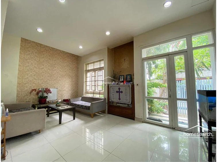This living area features a bright, open layout with polished white tile flooring and recessed ceiling lighting. The space is accented by a textured, patterned feature wall and connects directly to the outdoors through large glass-paneled doors, allowing for excellent natural light.