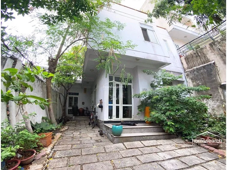 This exterior view shows a multi-story home with a stone-paved driveway and a lush, established garden. The architecture features clean, modern lines with large glass entry doors, and the property appears to be well-integrated into its natural, leafy surroundings.