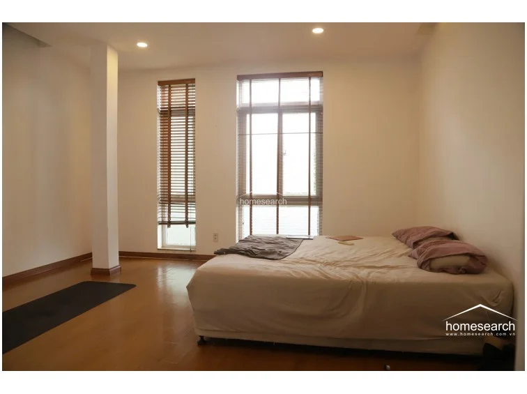 This bedroom features warm-toned wooden flooring and white walls, providing a minimalist and peaceful aesthetic. The room is illuminated by natural light from two windows fitted with wooden blinds and is currently furnished with a low-profile bed and a yoga mat. A structural column is visible, adding an architectural element to the otherwise simple and spacious room layout.