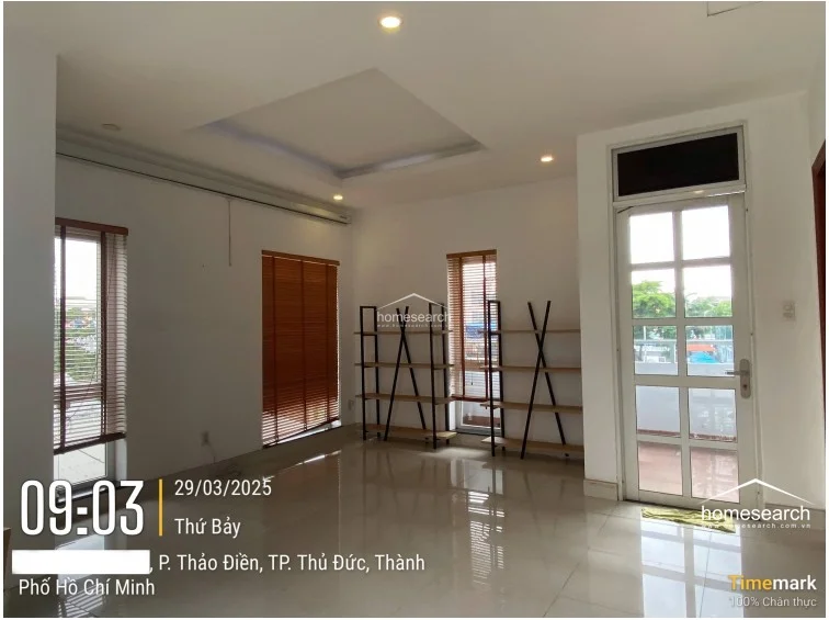 This room serves as a versatile living area or home office, characterized by high-gloss tile flooring and white walls that enhance the sense of space. It features multiple windows with wooden blinds and a glass-paned door that likely leads to a balcony or outdoor area, promoting good natural light and airflow. The room is sparsely furnished with two modern, open-frame shelving units, suggesting a flexible layout that could easily be adapted for various purposes.