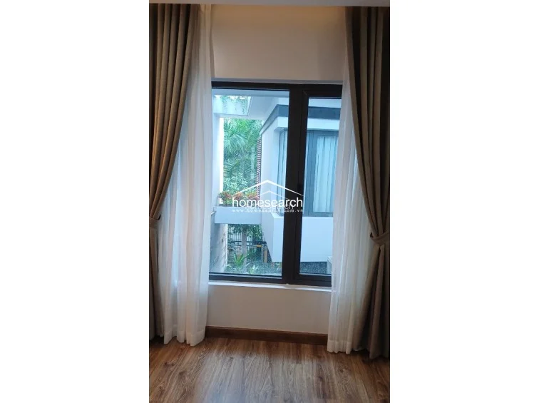 This bedroom features warm wood-look flooring and large windows dressed in layered beige and sheer curtains, allowing for ample natural light. The view looks out onto a neighboring building with lush greenery, creating a pleasant and private atmosphere.