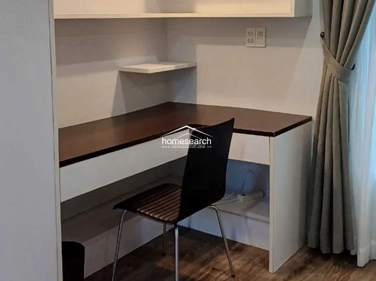 This dedicated workspace is integrated into the room with a custom L-shaped desk featuring a dark wood top and white base. Built-in shelving above the desk provides convenient storage, while the adjacent window with neutral curtains ensures a bright and productive environment.