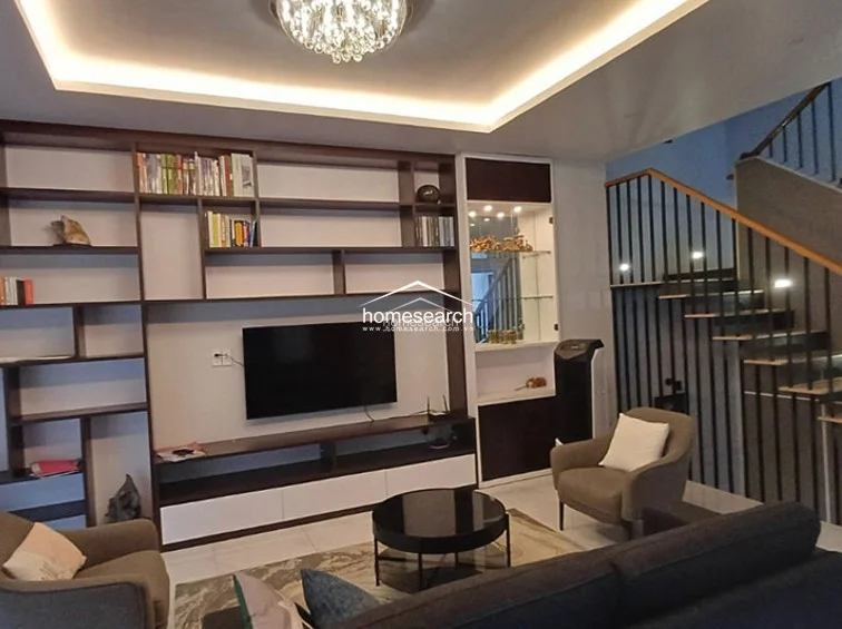This sophisticated living room centers around a custom, floor-to-ceiling wooden media console and bookshelf unit with integrated display lighting. The space is elegantly furnished with a dark sofa and armchair, set against light-colored flooring and a recessed ceiling with warm ambient cove lighting.