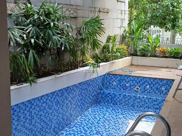This private outdoor space features a compact, rectangular swimming pool finished with vibrant blue mosaic tiles and integrated steps. The area is bordered by a wooden deck and a stone-clad wall, accented by lush tropical greenery that provides a natural, serene atmosphere.