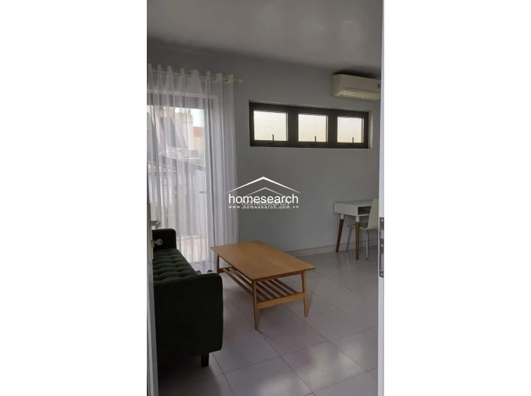 This functional living or study room features light grey walls, white tile flooring, and a large glass door leading to a balcony, ensuring excellent natural light. The room is equipped with a wall-mounted air conditioning unit, a green tufted sofa, a wooden coffee table, and a dedicated desk area, presenting a well-maintained and versatile space.