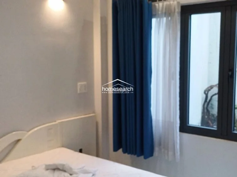 This bedroom features a clean, minimalist design with a focus on natural light provided by a large window dressed with blue curtains. The room is finished with light-colored walls and appears to be in well-maintained, move-in condition.