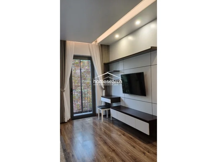 This living or bedroom area includes a modern media wall with integrated shelving and a desk, showcasing a sleek wood-and-white color scheme. The space features warm wood-look flooring and a glass door that opens to an outdoor area, providing excellent flow and natural illumination.