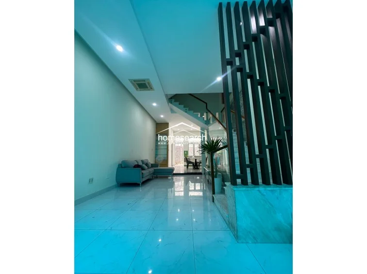 This open-concept living area features polished white marble-look tile flooring and a modern, minimalist aesthetic. A dark, vertical slat room divider creates a stylish transition between the seating area and the staircase, while recessed lighting and a ceiling-mounted AC unit maintain a clean, uncluttered look.