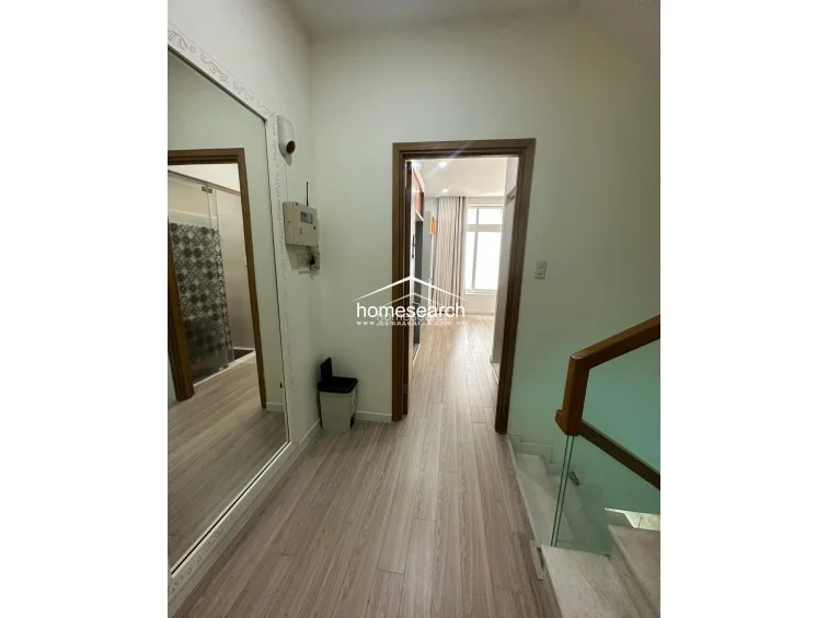This transition space serves as a hallway connecting a bedroom to a bathroom, featuring warm wood-look laminate flooring that contrasts with the neutral wall tones. A large, floor-to-ceiling wall mirror adds depth to the area, and the space includes a glass-railed staircase and a doorway leading to a well-lit bedroom.
