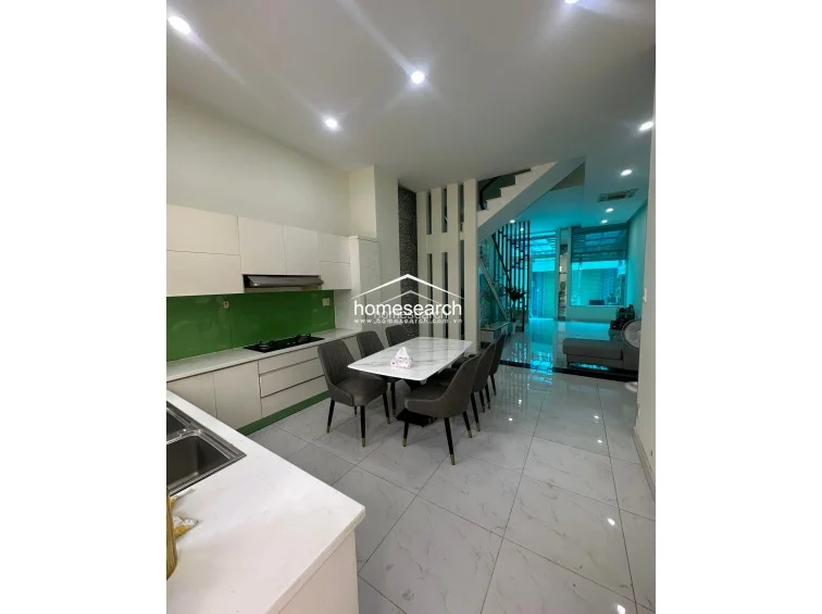 This modern kitchen and dining area is designed with a sleek, minimalist color palette, featuring white cabinetry, a vibrant green glass backsplash, and a white marble-topped dining table. The layout is highly functional, flowing seamlessly from the cooking zone to the living area, and is illuminated by multiple recessed ceiling lights.
