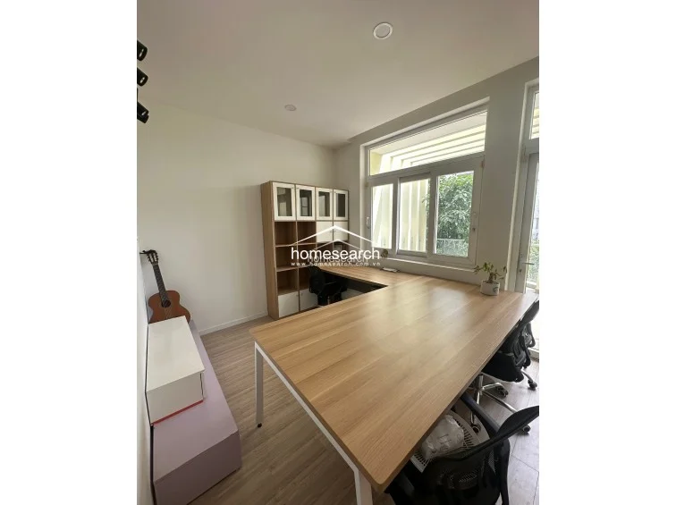 This home office or study room is bathed in natural light from a large window overlooking greenery, creating a bright and productive atmosphere. The room is efficiently furnished with a spacious L-shaped wooden desk and a tall, multi-compartment storage unit, all set against a neutral backdrop with light wood-tone flooring.