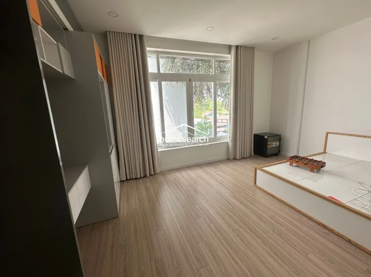This bedroom layout features a platform bed and a sleek, multi-functional storage unit with grey and orange accents. The room is designed for simplicity and light, utilizing full-length curtains and recessed lighting to enhance the modern, uncluttered atmosphere.