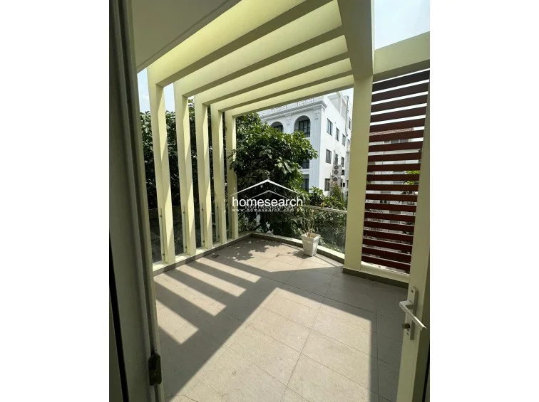 This modern balcony features a sleek pergola structure that provides architectural interest and partial shade. The space is finished with neutral floor tiles and a glass railing, offering a clean, open-air view of the surrounding neighborhood greenery.