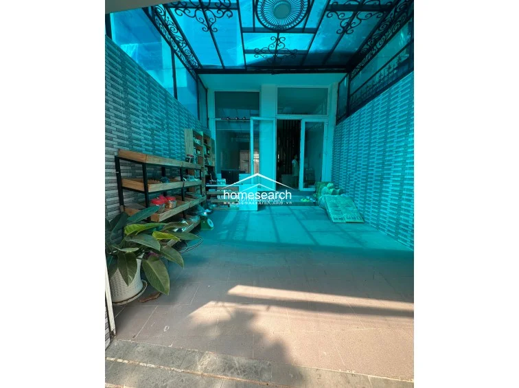 This covered outdoor area serves as a versatile utility or storage space, protected by a blue-tinted translucent roof. It features tiled walls and flooring, with integrated shelving units providing practical storage solutions for household items.