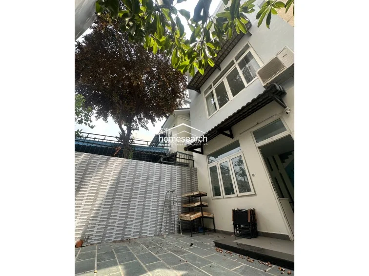 This exterior view shows a private courtyard or patio area paved with natural stone tiles, set against a backdrop of mature trees. The multi-story building features white exterior walls, large windows, and an external AC unit, reflecting a well-maintained property.
