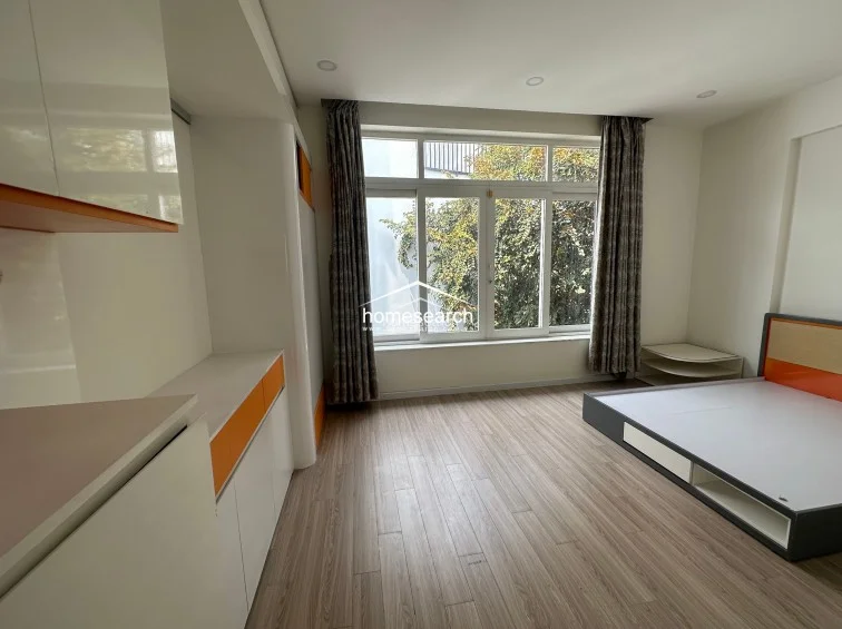 This bedroom showcases a modern, minimalist design with light wood-look flooring and a custom platform bed with integrated storage. A large window with floor-to-ceiling curtains allows for ample natural light, while built-in cabinetry with orange accents provides a cohesive and functional storage solution.