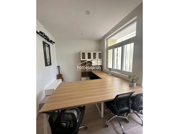 This functional home office is equipped with a large, L-shaped wooden desk and a matching multi-shelf storage unit. The space is well-lit by a wide window, creating an efficient workspace with a clean, professional aesthetic.