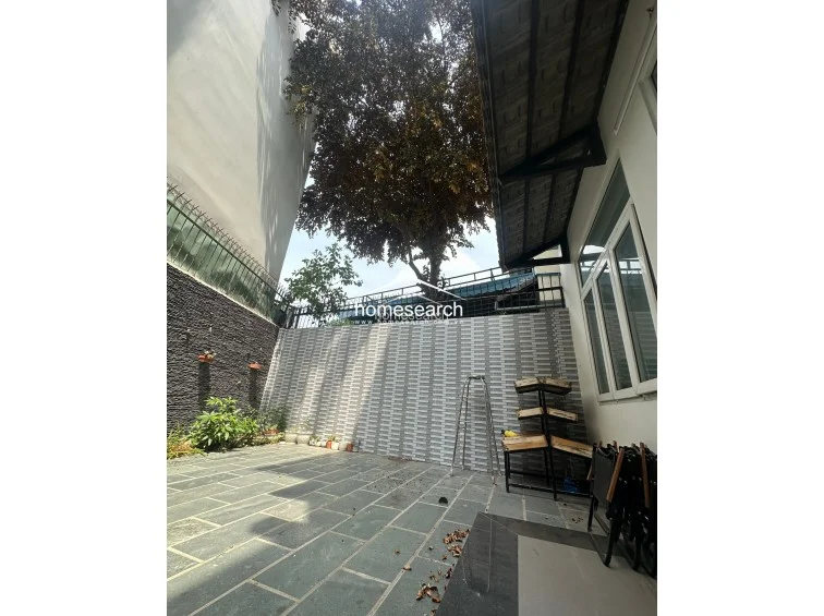 This outdoor terrace area features durable stone tile flooring and a combination of decorative textured wall finishes, providing a low-maintenance space for relaxation. The area is partially sheltered by an overhanging roof structure and benefits from natural shade provided by a large, mature tree overhead. Large windows connect the exterior space directly to the interior of the home, promoting a seamless indoor-outdoor flow.