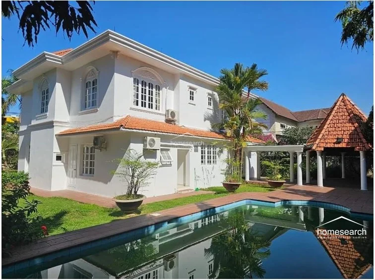 This exterior view showcases a two-story villa with a classic white facade, terracotta roof tiles, and multiple external AC units mounted on the walls. The property features a private swimming pool, a lush green lawn, and a covered outdoor pavilion supported by white columns, suggesting a well-maintained, resort-style living environment.