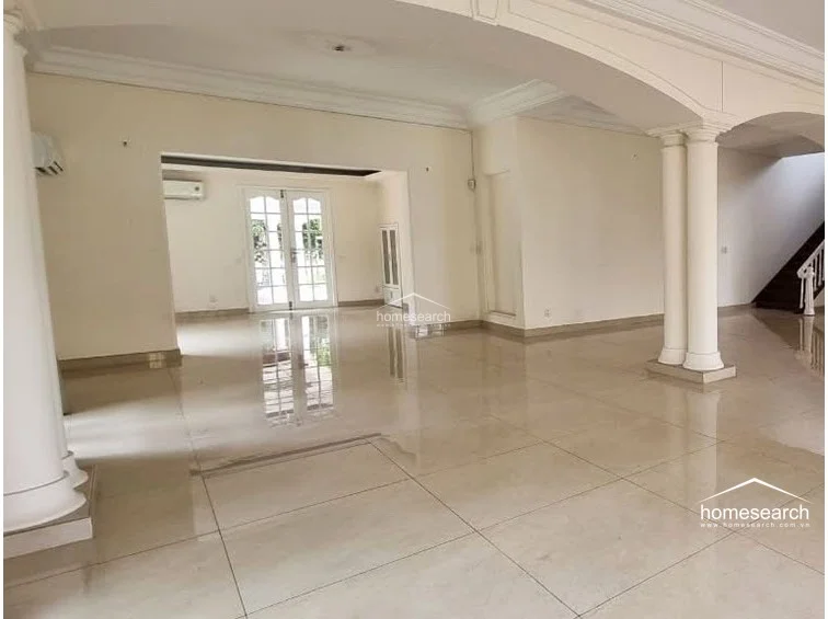 This spacious, open-concept living area features elegant architectural details, including grand columns and arched transitions that define the flow between rooms. The space is finished with high-gloss cream tile flooring and a neutral color scheme, complemented by wall-mounted air conditioning units and classic French doors that allow for ample natural light.