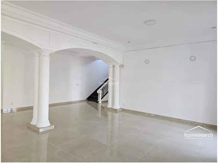 This view highlights the expansive, airy layout of the living space, characterized by high ceilings with decorative crown molding and a clean, minimalist aesthetic. The room is anchored by substantial white columns and leads toward a staircase, creating a sophisticated and well-maintained transition between the ground floor levels.