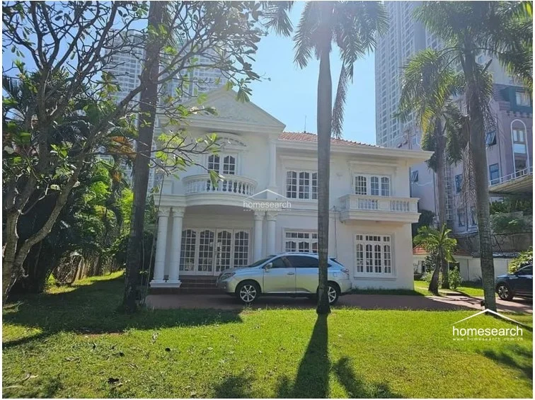 This image highlights the grand front entrance of the villa, which features a prominent balcony with balustrades and elegant arched windows. The property is set back from the road with a spacious, manicured lawn and a paved driveway, reflecting a high level of architectural detail and curb appeal.