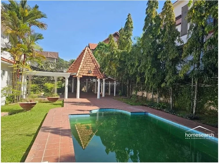 This angle emphasizes the outdoor leisure space, focusing on the swimming pool area and the adjacent shaded gazebo. The integration of mature trees, terracotta paving, and white structural columns creates a serene, private garden atmosphere suitable for outdoor entertaining.