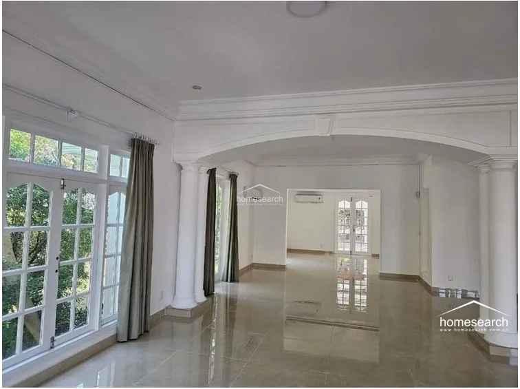 This perspective showcases the bright, sun-drenched living room, which is framed by large windows draped in neutral curtains and accented by classic architectural columns. The open floor plan encourages a natural flow, with air conditioning units installed for climate control and a clean, white-toned finish that enhances the sense of space.