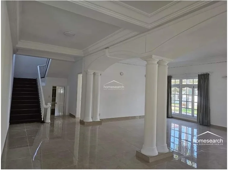 The living area maintains a consistent design language with its bright, neutral walls and polished tile flooring, offering a seamless connection to the outdoor greenery visible through the large, multi-paned windows. The architectural archways and columns provide a sense of structure and grandeur, while the overall condition appears pristine and ready for personalization.