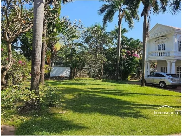 This perspective captures the expansive front yard, showcasing the villa's neoclassical design elements and its harmonious integration with the surrounding greenery. The wide lawn and mature palm trees provide a sense of privacy and tranquility, highlighting the property's generous plot size.
