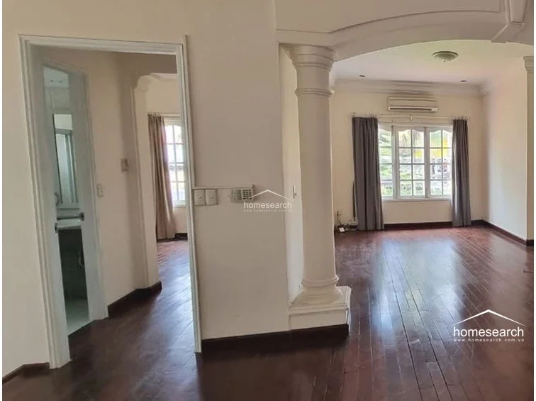 This transition area features a shift to warm, rich wood flooring, contrasting with the white walls and classical columns that define the main living space. A doorway leads to a compact, tiled bathroom, while the layout provides a clear, functional connection between the common living areas and private quarters.