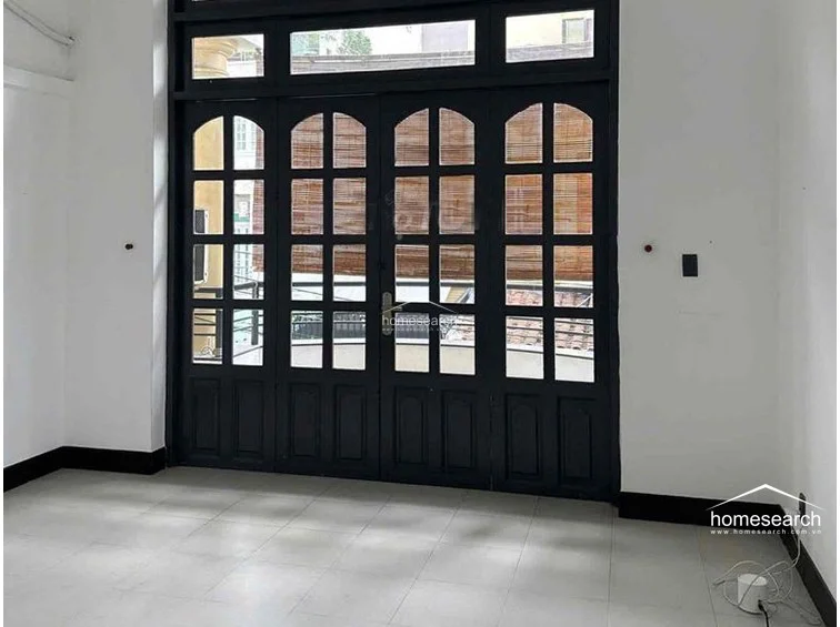 This room serves as a bright living or bedroom area, characterized by large, black-framed French doors that lead out to a balcony, allowing for significant natural light. The space maintains a minimalist aesthetic with white walls, dark baseboards, and neutral floor tiles, offering a versatile layout.